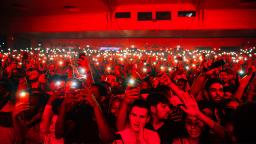 crowd in a music venue all holding their phones lights in the air, bathed in a red light.